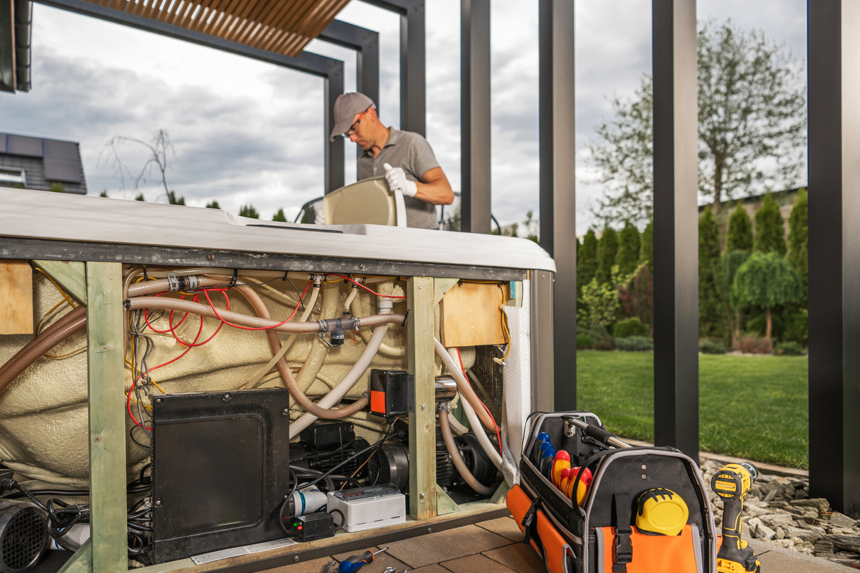 Technician inspecting exposed hot tub wiring and internal electrical components during outdoor installation under a backyard pergola.