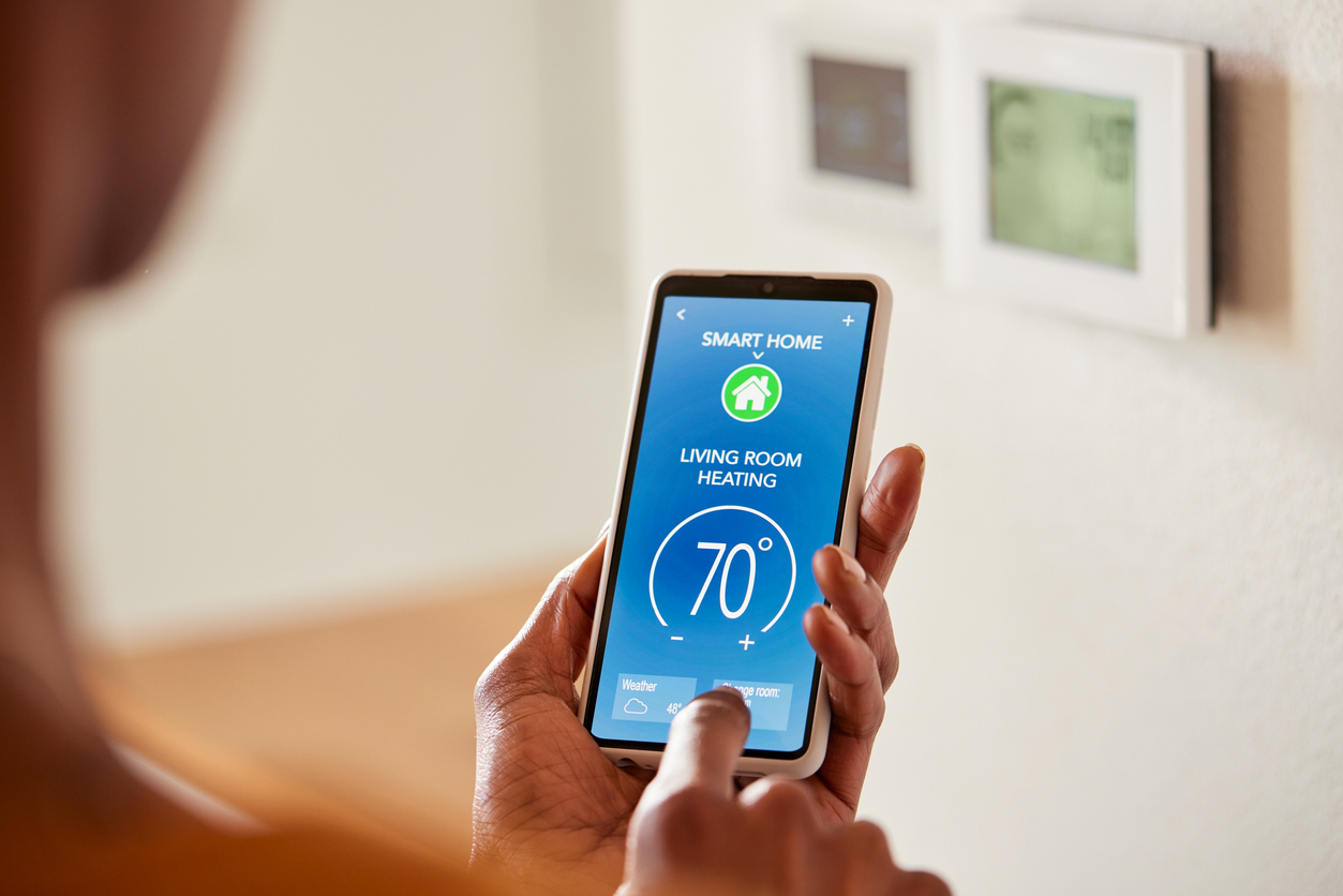 A person using a smart home app on their phone to adjust the temperature, with a smart thermostat mounted on the wall in the background.